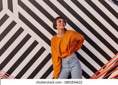 Positive woman with brunette hairstyle in orange sweatshirt and jeans smiling on striped backdrop. Cool lady with pink sunglasses posing outside.. Stock-foto