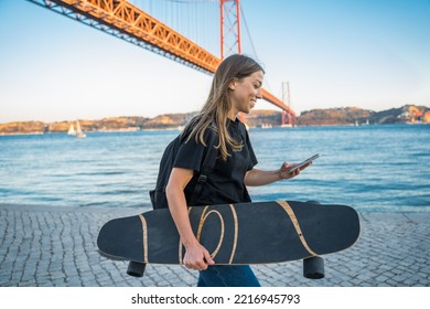 Positive Woman Carrying Skateboard And Chatting Smartphone On Go