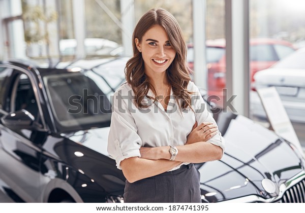 Positive woman with crossed arms smiling and\
looking at camera while standing near new vehicle during work in\
car dealership