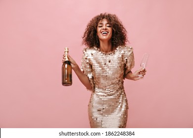 Positive woman with curly fluffy hairstyle in shiny modern clothes laughing and holding bottle with wine and glass on pink backdrop..
