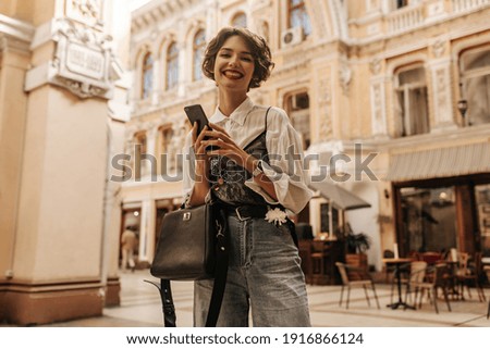 Positive woman with curly hair in jeans and white shirt smiling st street. Brunette girl with black handbag holds phone in city..