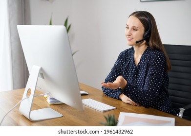 Positive Woman Is Customer Support Agent Sitting At The Desk And Consults Client Online, A Female Employee Wearing Headset Talking With Colleagues On The Distance, Involved Video Meeting