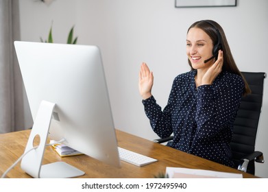 Positive Woman Is Customer Support Agent Sitting At The Desk And Consults Client Online, A Female Employee Wearing Headset Talking With Colleagues On The Distance, Involved Video Meeting