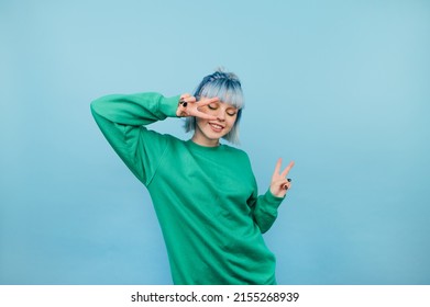 Positive woman in green sweatshirt and with colored hair dances on a blue background with a smile on her face. - Shutterstock ID 2155268939