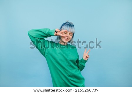 Positive woman in green sweatshirt and with colored hair dances on a blue background with a smile on her face.