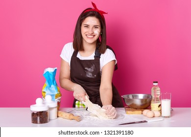 Positive Woman Has Dirty Face With Flour, Kneads Dough, Preapres Homemade Pastry During Weekend, Dressed In White T Shirt And Apron, Uses Diefferent Ingredients. Culinary And Baking Concept.