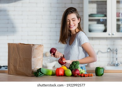 Positive Woman Holding Juicy Apples Near Paper Bag And Vegetables On Kitchen Table