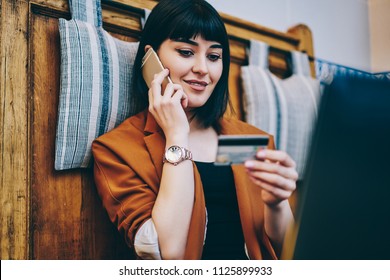 Positive Woman Looking At Number On Credit Card And Confirm Purchase Via Telephone Call To Customer Service, Smiling Hipster Girl Making Payment Via Smartphone Conversation While Resting Indoors