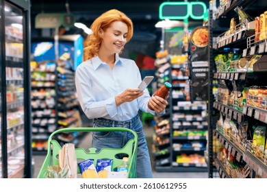 Positive woman scanning qr code of product, holding glass jar and using smartphone, standing near shelves. Concept of shopping and consumerism.