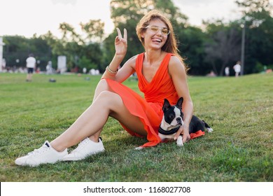 Positive Woman Sitting On Grass In Summer Park, Holding Boston Terrier Dog, Funny Face Expression, Wearing Orange Dress, Trendy Style, Slim Legs, Sneakers, Playing With Pet