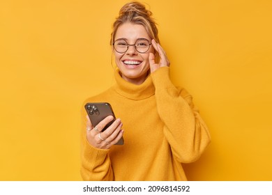 Positive woman smiles happily keeps hand on rim of spectacles feels cheerful wears comfortable sweater downloads cool application edits pics poses against yellow background. Technology concept - Shutterstock ID 2096814592