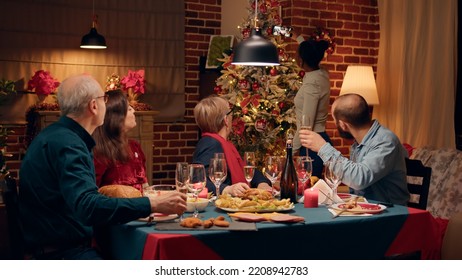 Positive Woman Standing Beside Christmas Tree While Taking Family Group Selfie Photo At Dinner. Festive Relatives Gathered Around Dinner Table While Celebrating Winter Holiday Together.