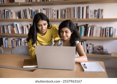 Positive young adult Indian students working on research study, educational project at laptop together, talking at computer with bookshelves in background, watching online content on screen - Powered by Shutterstock