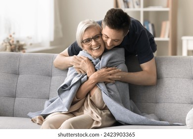 Positive young adult son man caring for happy elderly mature mother sitting on comfortable sofa, throwing warm plaid, scarf on moms shoulders, hugging from behind with love, affection - Powered by Shutterstock