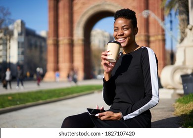 Positive young African American female with toothy smile wearing casual outfit holding mobile phone and drinking cup of takeaway coffee while resting in park in Barcelona on sunny day