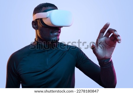 Positive young African American male millennial with dark hair and beard touching invisible screen and smiling while exploring cyberspace in VR goggles in studio