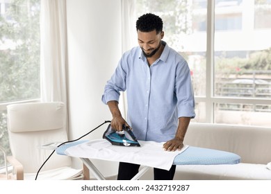 Positive young African American man ironing fabric in living room, pressing hot iron to board, using electrical equipment, doing housework, household chores, domestic activities in home apartment - Powered by Shutterstock