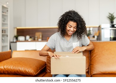 Positive young African American woman sitting on the couch with carton box on laps, feeling curious about ordered item from an online store, smiling client satisfied with fast courier delivery service