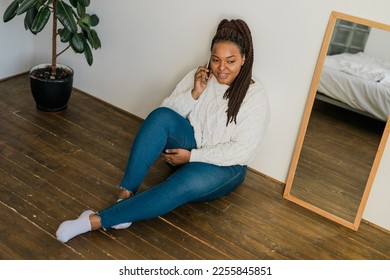 Positive young african american woman chatting on cellphone and smiling on floor at home. Cheerful young woman communicating with someone on smartphone indoors - Shutterstock ID 2255845851