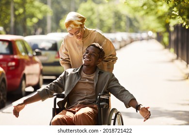 Positive young African-American disabled man chatting with friend who pushing him in wheelchair along street - Powered by Shutterstock