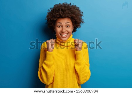 Positive young Afro American woman anticipates for announcement of results, smiles broadly and wears yellow jumper, triumphs joyfully, isolated over blue background. Victory celebration concept