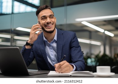 Positive Young Arab Guy In Suit Businessman Working At Modern Office, Sitting At Worktable In Front Of Laptop, Talking On Mobile Phone And Taking Notes, Looking At Copy Space And Smiling