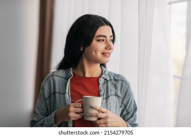 Positive young Arab woman drinking hot coffee, looking out window at home, enjoying peaceful morning. Charming Middle Eastern woman holding cup of warm beverage indoors