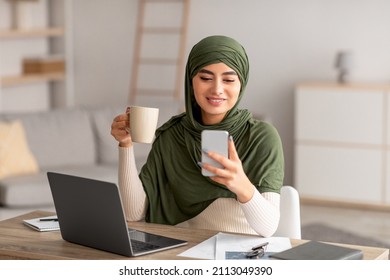 Positive young Arab woman in hijab using laptop and smartphone for distance job, drinking coffee while communicating online from home. Middle Eastern female speaking to colleagues remotely indoors