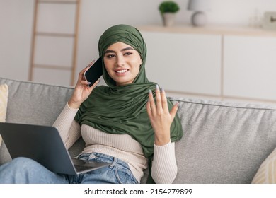 Positive Young Arab Woman In Hijab Using Laptop And Speaking On Smartphone At Home, Copy Space. Cheerful Middle Eastern Female Working On Pc, Speaking To Colleagues On Mobile Device