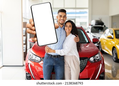 Positive Young Arabic Couple In Casual Standing By Brand New Red Car In Showroom, Showing Cell Phone With White Empty Screen, Mockup, Copy Space. Auto Dealership Online Concept