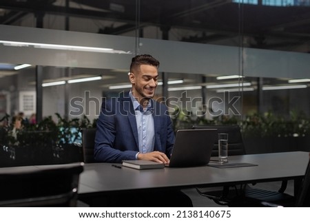 Positive young arabic manager working on laptop at modern hi-tech office, sitting at table, typing on computer keyboard, sending emails, communicating with business partners, copy space