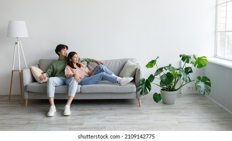 Positive Young Asian Couple Relaxing On Sofa, Cuddling And Looking Out Window In Living Room, Free Space. Millennial Husband And Wife Chilling On Comfy Couch, Spending Time Together
