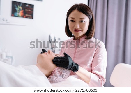 Positive young Asian female beauty master in black latex gloves smiling and looking at camera while doing massage on face of client during procedure