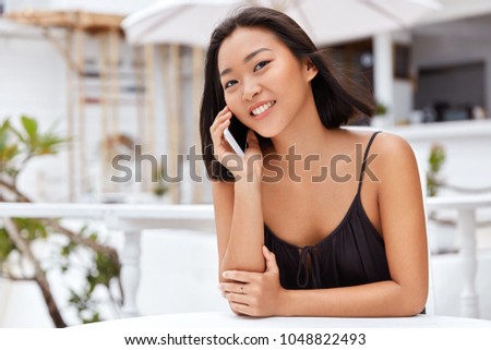 Positive young Asian female enjoys conversation via cell phone, shares her impressions about summer vacations with relatives, uses free roaming or call traffic, sits against coffee shop interior.