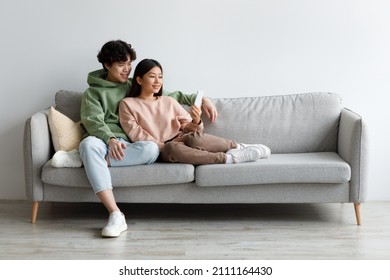 Positive Young Asian Husband And Wife Looking At Smartphone Screen, Sitting On Couch At Home, Copy Space. Millennial Couple Browsing Social Media, Chatting Online On Mobile Phone Indoors