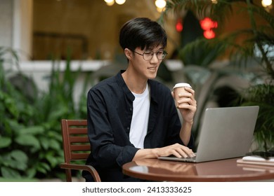 A positive young Asian man freelancer or digital nomad working remotely at a coffee shop, sipping coffee and working on his laptop computer. Urban city life concept - Powered by Shutterstock