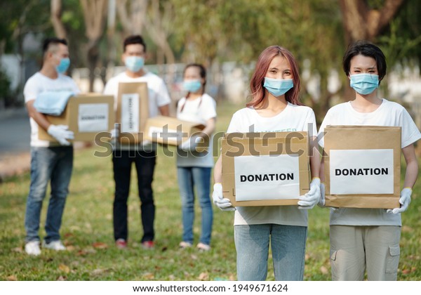 Positive young Asian volunteers in medical masks\
holding cardboard boxes of food and clothes for people who lost\
their job due to coronavirus\
pandemic