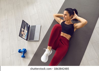 Positive young athlete exercising abs at home watching video workout lesson on laptop computer. Happy woman doing crunches following instructions of an online sports trainer. From above, high angle - Shutterstock ID 1863842485