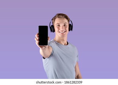 Positive Young Athlete In Headphones Showing Cellphone With Blank Screen Over Violet Studio Background, Mockup For Design. Fit Sportsman Listening To Music And Holding Phone With Space For Mobile App