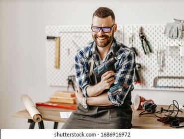 Positive Young Bearded Man In Apron And Eyeglasses Looking At Camera And Smiling While Standing With Drill In Hand In Modern Workshop
