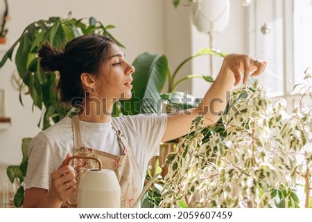 Positive young beautiful woman in apron is watering houseplants at home.Home gardening.Hobby concept.Biophilia design and urban jungle concept.Selective focus.