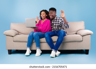 Positive Young Black Couple Having Web Call Or Video Chat On Laptop, Waving At Camera, Sitting On Couch On Blue Studio Background, Full Length. Remote Communication Concept