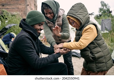 Positive young Black man in hat playing with kids against abandoned building and hanging clothes on rope in refugee camp