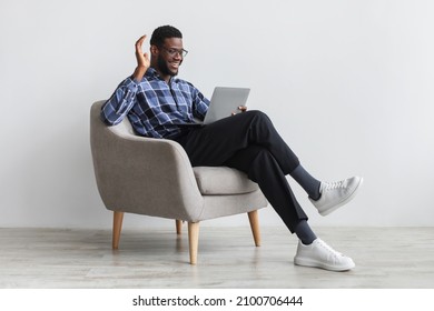 Positive young black man having web call or video conference on laptop, waving at camera, sitting in armchair against white studio wall, full length. Remote communication concept - Powered by Shutterstock