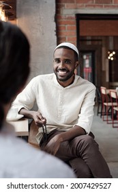 Positive Young Black Man In Kufi Cap Sitting At Table In Loft Cafe And Talking To Friend