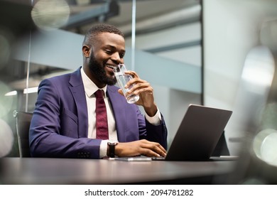 Positive young black man manager drinking water while working on laptop, typing on computer keyboard, making marketing research or sending emails, office interior, copy space