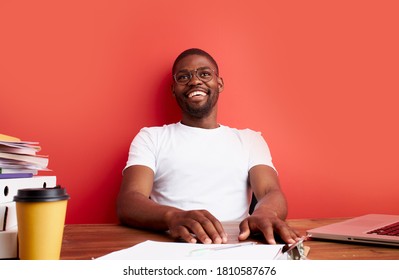 positive young black man sit at office desk, afro american guy smile at camera, enjoy work, isolated red background