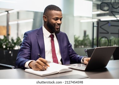 Positive Young Black Manager In Suit Sitting At Workdesk, Typing On Laptop Keyboard, Taking Notes, African American Businessman Attending Business Webinar Online, Copy Space. Business Education