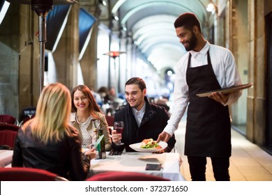 Positive Young Black Waiter Serving Terrace Restaurant Guests At Table.Focus On The Man