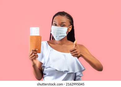 Positive Young Black Woman Wearing Protective Face Mask, Holding Passport And Plane Tickets, Showung Thumb Up Gesture On Pink Studio Background. Traveling Abroad During Covid Outbreak